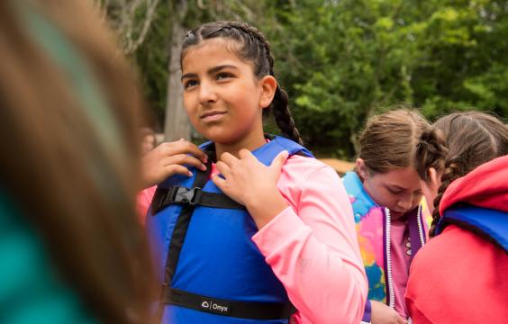 A young girl wearing a life jacket. Others in the background are putting life jackets on.