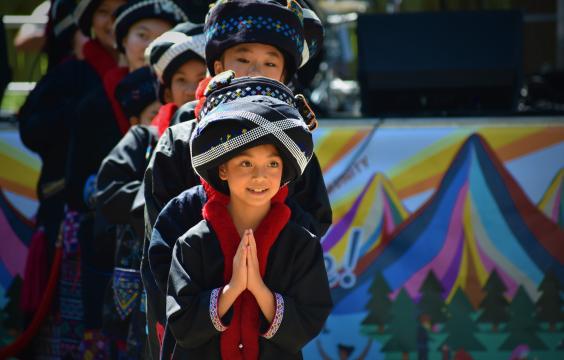 A group of young Mien dancers dress in traditional clothing. The girl in the front smiles and holds up her hands in praying gesture. 